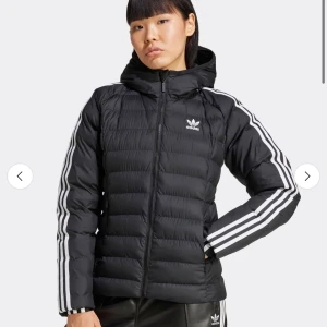 Svart Adidas pufferjacka med huva - Svart pufferjacka från Adidas med ikoniska vita ränder längs ärmarna och Adidas-logga på bröstet. Jackan har huva, dragkedja framtill och quiltad design. Perfekt för dig som gillar sportig och clean stil.
