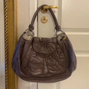 Grå handväska från Miu Miu i skinn - Snygg grå handväska från Miu Miu i mjukt skinn med silverfärgade metalldetaljer och dubbla handtag. Väskan har ett veckat lock framtill, innerfack med dragkedja. . Perfekt för dig som vill ha en lyxig och rymlig väska. Höjd utsn handtag 32 cm och bredden på det bredaste stället 48 cm.  Äkta / autentisk. 