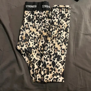 Leopardmönstrade leggings från Stronger - Snygga leggings från Stronger med leopardmönster i beige, svart och bruna toner. De har en bred svart midja med loggan STRONGER tryckt framtill. Perfekta för dig som vill sticka ut på gymmet eller i vardagen.