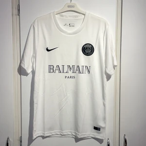 Vit PSG x Balmain fotbollströja Nike M - Snygg vit fotbollströja från Nike i samarbete med PSG och Balmain. Tröjan har korta ärmar, rund hals och är gjord i lätt Dri-Fit material som andas. PSG-logga och Balmain Paris tryck på bröstet, samt svart Nike Swoosh. Perfekt för dig som gillar exklusiva samarbeten.
