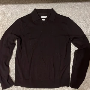 Svart merinotröja från These Glory Days - Mörkbrun långärmad tröja från These Glory Days i mjuk merinoull. Klassisk krage och v-ringad hals ger en clean och stilren look. Passformen är normal och materialet känns lyxigt och skönt mot huden. Storlek M och nästan aldrig använt. 