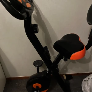 Klarfit X-Bike 700 motionscykel - Klarfit X-Bike 700 träningscykel med digital display, justerbart motstånd och bekväm sadel. Svart och orange design, kompakt och hopfällbar modell. Cykeln ser ut att vara i gott skick utan synligt slitage eller defekter. Perfekt för hemmaträning och kondition.
