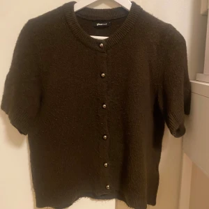 Brun kortärmad kofta från Gina Tricot - Mysig brun kofta från Gina Tricot med korta ärmar och rund hals. Koftan har ribbade kanter och snygga knappar framtill. Perfekt att slänga över en topp för en chill look. Materialet känns mjukt och lite fluffigt.