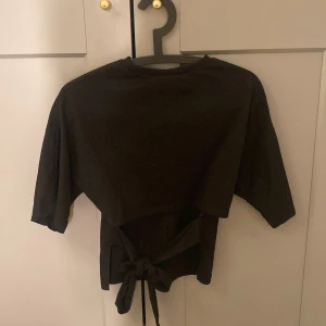 Svart croppad topp med knyt från Nelly - Svart croppad topp från Nelly i storlek XXS. Toppen har korta ärmar och en snygg knytning vid ryggen. Materialet känns mjukt och lätt, perfekt för dig som gillar stilrena och enkla plagg med en twist.