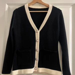 Elegant svart kofta  - Svart kofta med krämvita kanter och eleganta pärlknappar framtill. Koftan har två fickor och en v-ringad hals. Materialet är stickat och känns mjukt. Från SHEIN