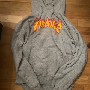 Grå Thrasher hoodie med flames - Säljer en grå hoodie från Thrasher med ikoniskt flames-tryck i gult och rött över bröstet. Tröjan har huva med snörning och stor magficka framtill. Materialet är mjuk bomull och polyester, perfekt för chill och skate. Passformen är normal och ärmarna är långa.