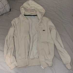 Beige hoodie från Lacoste med dragkedja KVITTO FINNS - Snygg beige hoodie från Lacoste med klassisk krokodillogga på bröstet. Tröjan har huva med snörning, två fickor fram och hel dragkedja med Lacoste-detalj. Mjuk insida och ribbade muddar vid ärmslut och nederkant. Perfekt för chill dagar.