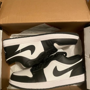 Nike Air Jordan 1 Low svart/vit (panda) - Oanvända Nike Air Jordan 1 Low i svart och vitt med klassisk Swoosh på sidan och Air Jordan-logga på hälen. Skorna har snörning, rund tå och platt sula. Tillverkade i skinn med perforerad tå för extra ventilation. Perfekt för dig som gillar streetstyle och sneakers med ikonisk design fick i present inte min still därför den nu är till salu .Det är bara att prutta på 