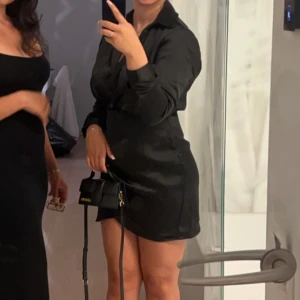 Svart omlottklänning med lång ärm - Snygg svart kort omlottklänning med lång ärm och v-ringad krage. Klänningen har en figurnära passform och är tillverkad i ett glansigt satinliknande material som ger en lyxig känsla. Perfekt för dig som gillar stilrena och eleganta plagg.