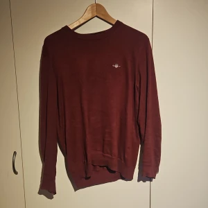 Vinröd stickad tröja från Gant - Vinröd stickad tröja från Gant med rund hals och broderad logga på bröstet. Tröjan är i herrstorlek small och som syns på bild så har tröjan en liten lagning i höger armhåla. Förutom det så är tröjan i bra skick och den fyller sitt syfte.