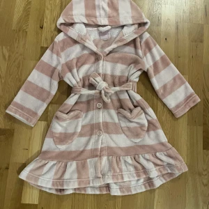Randig rosa och vit morgonrock  - Supermjuk morgonrock i fleece med breda rosa och vita ränder, huva, knappar framtill och knytskärp i midjan. Två hjärtformade fickor och volangkant nertill ger en gullig touch. Perfekt för mysiga dagar hemma.
