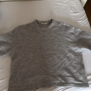 Grå stickad tröja från Gina Tricot - Mysig grå stickad tröja från Gina Tricot i storlek S. Tröjan har en klassisk rund halsringning och ribbade muddar vid ärmslut och nederkant. Modellen är lite loose och har kortare ärmar, perfekt för lager-på-lager under hösten och vintern.
