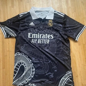 Real Madrid svart draktröja Adidas - Snygg svart Real Madrid fotbollströja från Adidas med vit krage och detaljer. Tröjan har ett stort drakmotiv över hela framsidan och baksidan, samt klubbmärke och sponsortryck på bröstet. Kortärmad modell i lätt och ventilerande material.