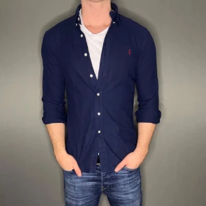 Ralph Lauren skjorta  - Mörkblå skjorta från Ralph Lauren med klassisk krage och knappar framtill. Skjortan har lång ärm och den ikoniska röda loggan broderad på bröstet. Tillverkad i mjuk bomull för en skön känsla och snygg passform.