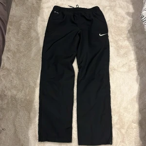 Nike Dri-Fit svarta träningsbyxor S - Svarta träningsbyxor från Nike med Dri-Fit-teknologi och broderad vit swoosh på benet. Byxorna har normal passform, elastisk midja med snörning och är tillverkade i lätt polyester med meshfoder för extra ventilation. Perfekta för sport och aktivitet.