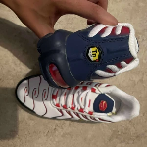 Nike Air Max Plus vita/röda sneakers - Nike Air Max Plus sneakers med vit mesh-överdel, röda och blå detaljer samt gula inslag på loggan. Snyggt vågigt mönster och synlig Air Max-dämpning i sulan. Platt sula och snörning framtill. Perfekt för dig som gillar sportig och unik stil.