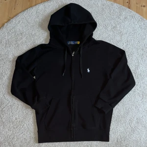 Svart hoodie från Polo Ralph Lauren - Svart hoodie från Polo Ralph Lauren med dragkedja och klassisk vit broderad logga på bröstet. Tröjan har huva med snörning, två fickor framtill och är tillverkad i mjuk bomull. Perfekt för en chill och stilren look. Har dock ganska stark doft av parfym. 