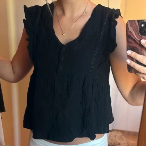 Svart blus med volang från J'adore - Svart blus med v-ringning och knappar framtill. Snygga volangdetaljer på axlarna!