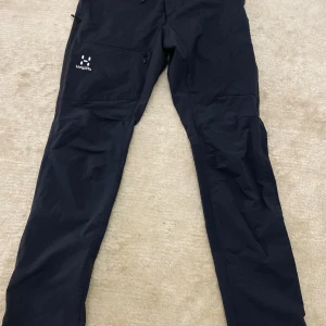 Svarta friluftsbyxor från Haglöfs - Svarta friluftsbyxor från Haglöfs i storlek 46. Byxorna har en rak passform, praktisk dragkedja på benfickan och är tillverkade i slitstarkt syntetmaterial. Perfekta för utomhusaktiviteter och har diskret Haglöfs-logga på benet.