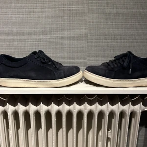 Mörkblå Sandö skor  - Snygga mörkblå sneakers i mocka med klassisk vit platt sula. Skorna har rund tå, svarta snören och stilren design som passar till det mesta. Perfekta för dig som gillar en clean och enkel look.