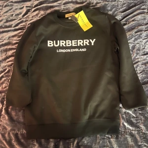 Svart sweatshirt från Burberry - Svart sweatshirt från Burberry med vit logotyptext 'BURBERRY LONDON ENGLAND' på bröstet. Tröjan har rund hals och långa ärmar, samt ribbade muddar vid ärmslut och nederkant. Perfekt för dig som gillar stilrena designer och ikoniska märken.