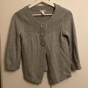 Trekvartsärmad kofta - Grå kofta från H&M med bred ribbad krage och tre stora knappar framtill. Koftan har trekvartsärm och snygga stickade detaljer längs sidorna. Perfekt för lager-på-lager och chill stil. Materialet är mjukt och känns som bomull.