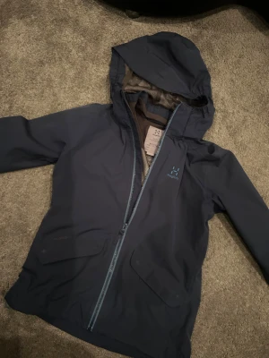 Mörkblå regnjacka från Haglöfs - Snygg mörkblå regnjacka från Haglöfs med huva och meshfoder. Jackan har två stora fickor framtill och hel dragkedja i matchande blå ton. Perfekt för blöta dagar och outdoor-aktiviteter. Stilren design med diskret Haglöfs-logga på bröstet.