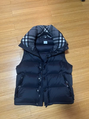 Burberry jacka - Svart pufferjacka från NAKED med avtagbara ärmar så du kan bära den som väst. Jackan har en stor grå rutig krage, dragkedja och knappar framtill samt två fickor med dragkedja. Perfekt för dig som gillar streetstyle och vill ha en jacka som sticker ut.