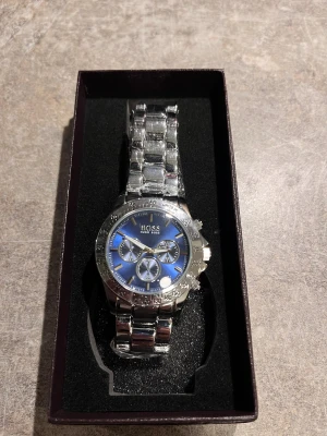 Blå och silver Hugo Boss klocka - Snygg kronografklocka från Hugo Boss med blå urtavla och silverfärgat armband i rostfritt stål. Klockan har tre mindre visartavlor, markerade index och detaljerad bezel. Perfekt accessoar för dig som gillar stilrena och moderna detaljer.