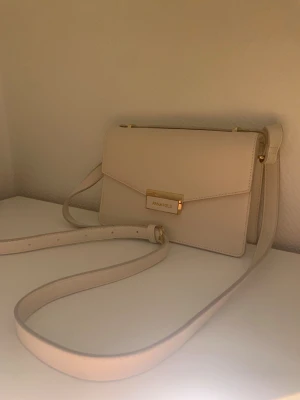 Beige axelväska från Anna Field - Snygg beige axelväska från Anna Field med stilren, fyrkantig form och guldfärgade detaljer. Väskan har justerbar axelrem och ett elegant spänne framtill med logga. Perfekt för dig som gillar minimalistisk och clean stil.