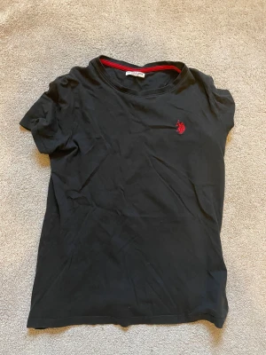 Svart t-shirt från U.S. Polo Assn - Svart t-shirt från U.S. Polo Assn med röd logga broderad på bröstet och röd detalj längs insidan av halsringningen. Klassisk rund hals och korta ärmar. Tillverkad i mjuk bomull som är skön mot huden.