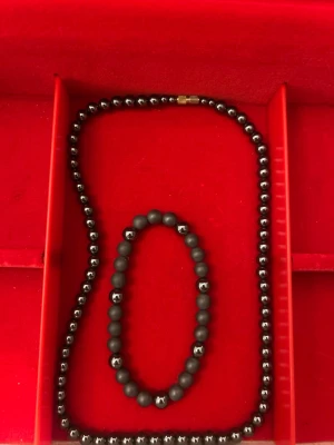 Svart pärlhalsband och armband - Snyggt set med svart pärlhalsband och matchande armband. Halsbandet har blanka, runda pärlor och ett diskret guldfärgat lås. Armbandet består av matta och blanka pärlor i svart, vilket ger en cool kontrast. Perfekt för dig som gillar stilrena accessoarer.