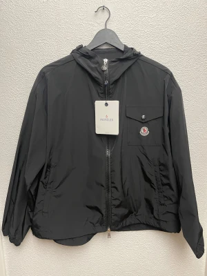Svart vindjacka från Moncler med huva - Svart vindjacka från Moncler med huva, dragkedja framtill och en bröstficka med knapp och broderad logotyp. Jackan har en patch med Moncler-logga på ärmen och justerbar dragsko i huvan. Tillverkad i lätt polyester, perfekt för blåsiga dagar.
