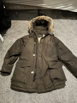 Woolrich Arctic Parka brun - Säljer en Woolrich Arctic Parka i mörkbrun färg med beige quiltat foder och pälskantad huva. Jackan har stora fickor framtill, dragkedja och knappar, samt långärmad design. Perfekt för kalla vinterdagar och riktigt snygg streetstil. Äkta päls går inte att ta bort pälsen. 