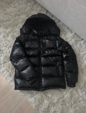 Svart pufferjacka Polo Ralph Lauren XS - Snygg svart pufferjacka från Polo Ralph Lauren med glansig finish, huva och dragkedjor. Jackan har klassisk röd broderad logga på bröstet och quiltad design. Perfekt för kalla dagar och riktigt street i stilen.