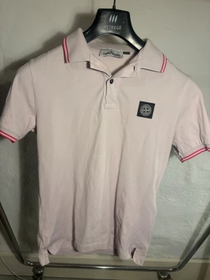 Stone Island Pike  - Rosa pikétröja från Stone Island med klassisk krage och röda ränder på krage och ärmslut. Tröjan har korta ärmar och en broderad logotyp på bröstet. Tillverkad i bomull för en skön känsla och snygg passform. Pris 249kr, storlek S.  Vid funderingar är det bara till att höra av dig 
