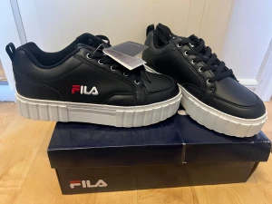 Svarta Fila sneakers med plattform - Svarta Fila sneakers med chunky vit plattformsula och snörning. Skorna har rund tå och Fila-logga broderad i vitt och rött på sidan och hälen. Ovandelen är i syntetmaterial med mjuk textil på insidan. Snygg kontrast mellan svart och vit, perfekt för dig som gillar streetstyle.