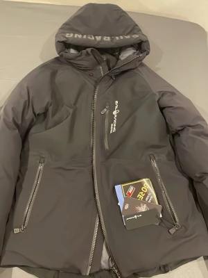 Svart dunjacka från Sail Racing - Svart dunjacka från Sail Racing med GORE-TEX-material, stor huva med logga och flera dragkedjeförsedda fickor. Insidan är quiltad och jackan har en clean, sportig look med Sail Racing-tryck på bröstet. Perfekt för kalla dagar och outdoor.