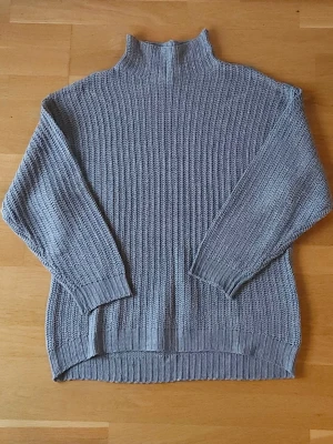 Grå stickad tröja - Mysig grå stickad tröja med hög krage och långa ärmar. Tröjan har en oversized passform och ribbade muddar vid ärmslut och nederkant. Perfekt för kyliga dagar och enkel att styla till jeans eller leggings.