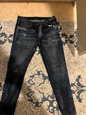 Dsquared2  jeans med färgstänk - Mörkblå Dsquared2  jeans med coola färgstänk och slitningar. Klassisk femficksmodell, rakt snitt och tydlig röd ICON-lapp bak i midjan. Jeansen har en smal passform och är tillverkade i robust denim för en edgy streetlook. 30/32