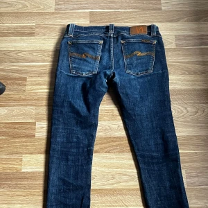 Blå jeans från Nudie Jeans - Säljer ett par snygga blå jeans från Nudie Jeans med klassisk femficksdesign och orange sömmar. Jeansen har en slitning vid grenen och en läderpatch med logga på baksidan. Perfekta för en avslappnad stil.