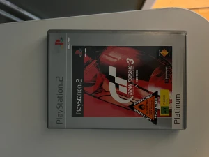 Gran Turismo 3: A-Spec (PlayStation 2 Platinum) - Gran Turismo 3: A-Spec till PlayStation 2 Platinum. Omslaget och fodralet ser ut att vara i gott skick med tydliga tryck och inga synliga skador. Perfekt för samlare eller racingentusiaster som vill uppleva klassisk bilsimulator på PS2.