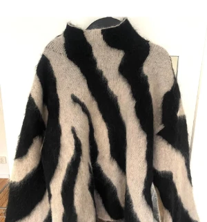 Beige och svart zebramönstrad stickad tröja - Säljer en fluffig stickad tröja med beige och svarta zebraränder. Tröjan har hög hals och långa ärmar, perfekt för dig som vill sticka ut med ett djuriskt mönster. Materialet är mjukt och ger en cozy vibe, passar dig som gillar statement-plagg.