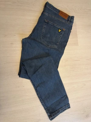 Blå jeans från Lyle & Scott W36 skinnyfit - Snygga blå jeans från Lyle & Scott med klassisk femficksdesign och gul broderad logga på bakfickan. Jeansen har en rak passform och är tillverkade i slitstarkt denimtyg. Perfekt för dig som gillar stilrena och tidlösa jeans.