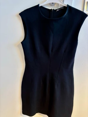 Svart klänning från Zara - En stilren svart klänning från Zara i figurnära modell. Klänningen är ärmlös, har rund halsringning och dragkedja bak. Storlek L (men sitter tight, så passar den som brukar ha storlek S/M). Längd. Ca 80 cm