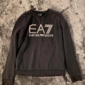 Svart EA7 Emporio Armani sweatshirt - Svart sweatshirt från EA7 Emporio Armani med stor logga framtill i vitt och grått. Tröjan har rund halsringning, långa ärmar och ribbade muddar. Perfekt för dig som gillar street hoodies, dock inte det bästa skicket eftersom att slutet på ärmarna har lösa trådar.