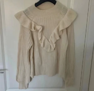 Beige stickad tröja med volang - Superfin beige stickad tröja med breda volanger över axlarna och framtill. Tröjan har ribbad struktur och långärmade ärmar med breda muddar. 