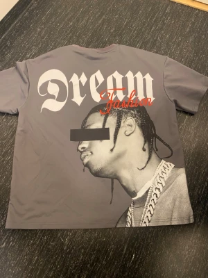 Travis Scott tröja  - Snygg grå t-shirt med stort Dream Fashion-tryck på ryggen och coolt motiv av en person med kedja Travis Scott Framsidan har en liten Dream-logga i vitt. T-shirten har rund hals och korta ärmar, perfekt för dig som gillar streetwear och statement-plagg.