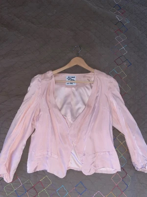 Ljusrosa satin kavaj från Just Female - Säljer en ljusrosa kavaj från Just Female i glansigt satinliknande material. Jackan har en öppen design utan knappar, långa ärmar och två dekorativa fickor framtill. Perfekt för dig som vill ha en chic och feminin look.