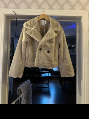Beige pälsjacka med stora slag - Säljer en supermjuk och fluffig beige pälsjacka från lager157 Jackan har stora slag, dubbelknäppning med svarta knappar och är croppad i modellen. Perfekt för dig som vill ha en trendig och mysig look i vinter. Aldrig använd med lappen kvar,  nypris 400